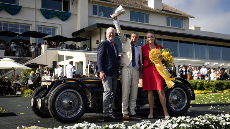 Bugatti Type 59 Sports 1934 року, який отримав найкращий приз на Pebble Beach Concours d'Elegance 2024, стоїть на подіумі перед будівлею, а його власник гордо тримає кубок перед ним