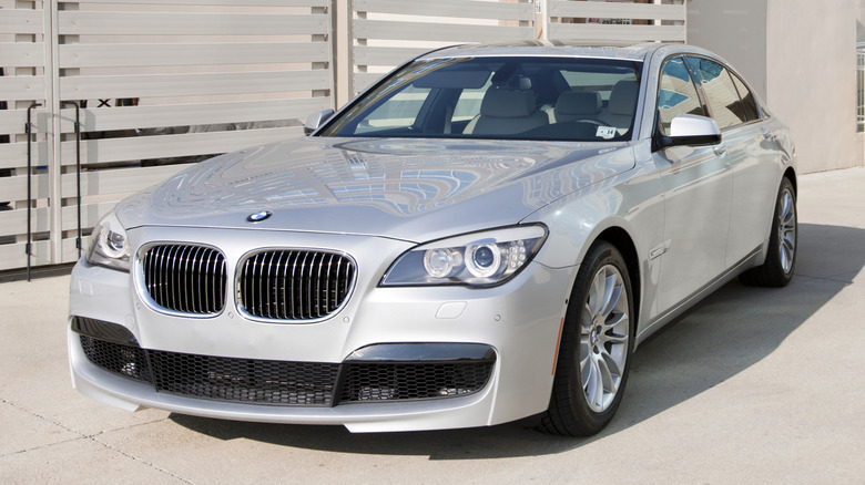 Сріблястий BMW 7 Серії 2012 року, припаркований біля паркану.
