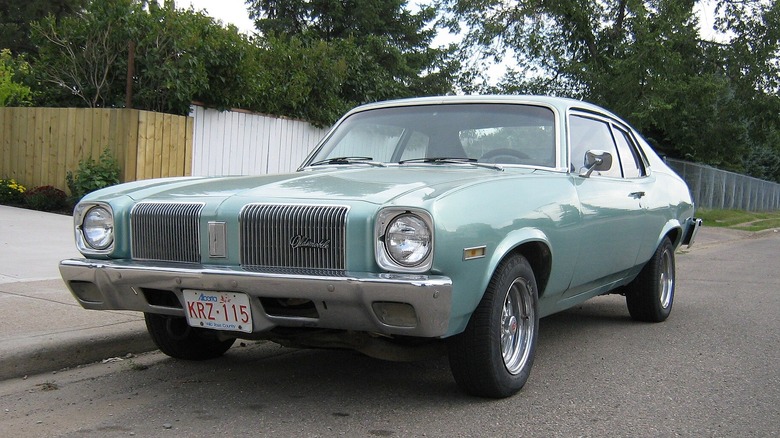 Світло-блакитний Oldsmobile Omega, припаркований на вулиці житлового кварталу з дерев'яною огорожею позаду.