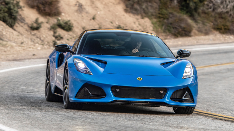 Фронтальний знімок синього Lotus Emira, що їде поворотом перед гірським схилом, вкритим чагарниками
