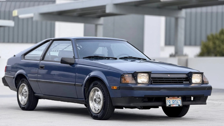 Синя Toyota Celica GT 1984 року в кузові хетчбек, припаркована на бетоні з увімкненими висувними фарами