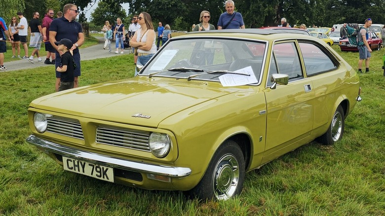 Гірчично-жовтий Morris Marina 1.3 SDL coupe 1971 року з хромованими ковпаками, припаркований на траві на відкритому автомобільному шоу.