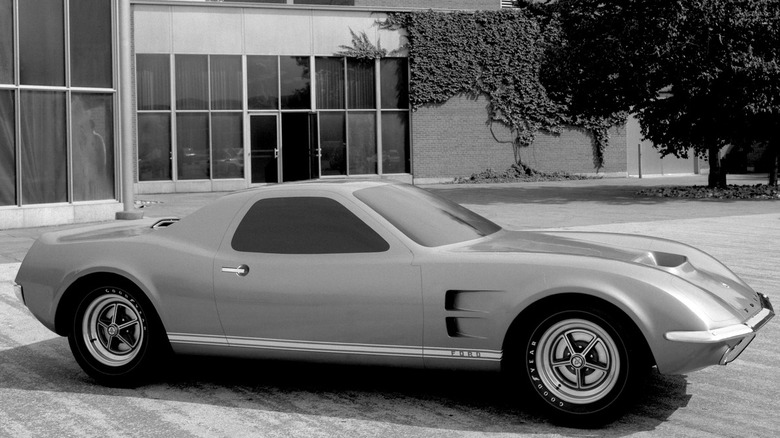 Чорно-біле фото концепту 1967 Ford Mach 2 з центральним розташуванням двигуна, припаркованого біля будівлі стилю Ford, що показує його довгу задню частину, вентиляційні отвори на передніх крилах і класичні диски в стилі магнієвих.
