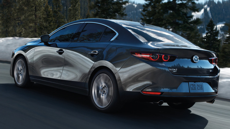 Сірий седан Mazda Mazda3 в русі по дорозі, повз дерева та засніжену землю.