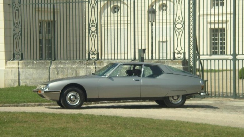 Елегантний сріблястий Citroën SM, розкішний гран-турінг, припаркований на гравії перед високими декоративними кованими воротами палацу.