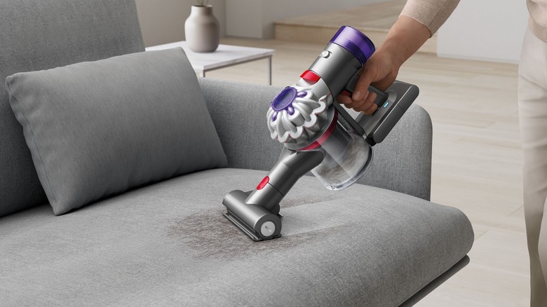 Ручний пилосос Dyson Car and Boat використовується для видалення шерсті з сірого дивана.