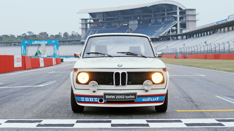 Білий BMW 2002 Turbo з додатковими фарами та написом 'turbo' задом наперед на передньому спойлері, що стоїть на стартово-фінішній лінії гоночної траси.