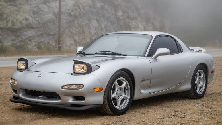 Передня тричетвертна частина сріблястого Mazda RX-7 FD, припаркованого на ґрунтовій дорозі перед туманним краєвидом скелястих гір із увімкненими фарами