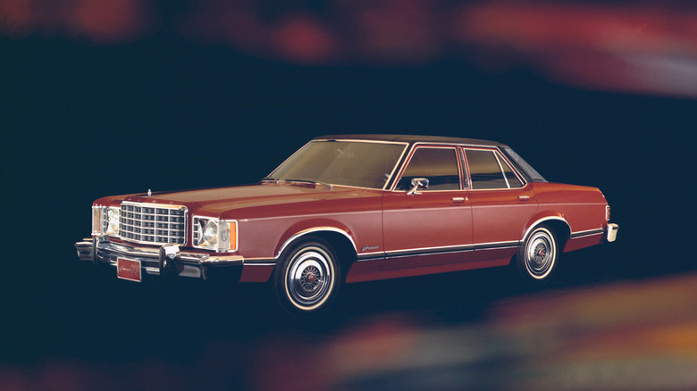 Червоний Ford Granada 1975 року, чотиридверний седан з чорним вініловим дахом, ковпаками на колесах і хромованою решіткою радіатора на темному, похмурому фоні.