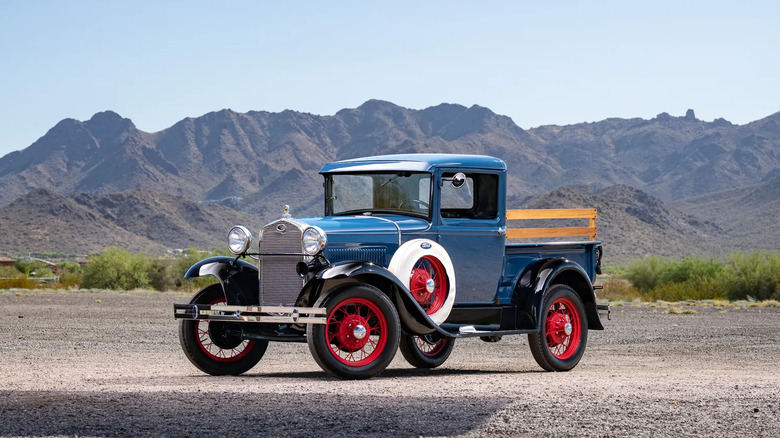 Синій пікап Ford Model A з червоними колесами, припаркований на ґрунті перед видом на гори та чисте небо