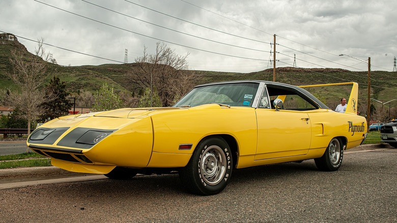 Яскраво-жовтий Plymouth Superbird 1970 року, що демонструє свій аеродинамічний гострокінцевий ніс, високе заднє антикрило та чорні графічні елементи на капоті, припаркований на асфальті.
