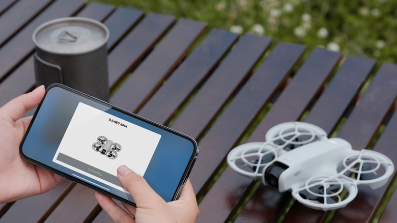 Людина керує дроном DJI Neo за допомогою програми на смартфоні, тоді як дрон і банка стоять поруч на столі з прорізами