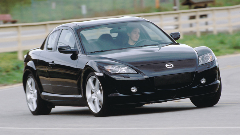 Чорна Mazda RX-8 2004 року, що проїжджає поворот перед травою та дерев'яним парканом