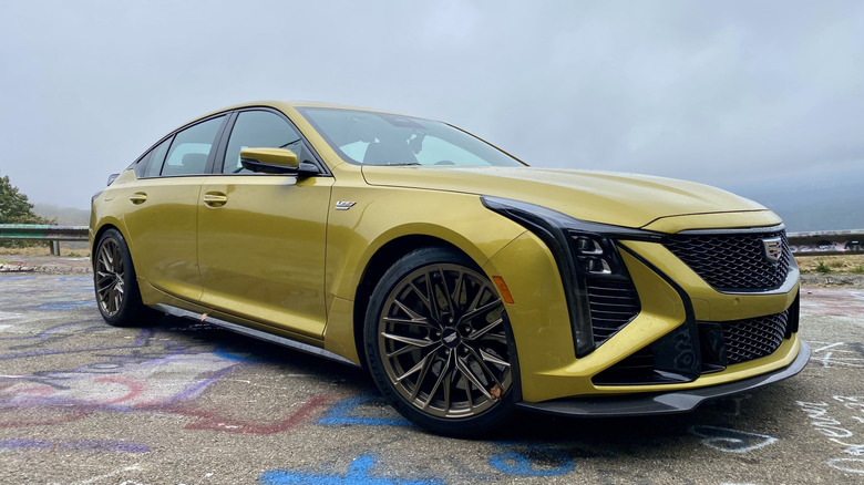 Передня тричетвертна частина жовтого Cadillac CT5-V Blackwing, припаркованого на асфальті, вкритому графіті, перед сірим хмарним небом