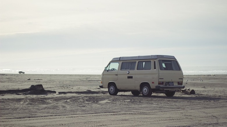 Блідо-жовтий Volkswagen Vanagon на пляжі в похмурий день з сонцем, що створює ефект вицвілого зображення.